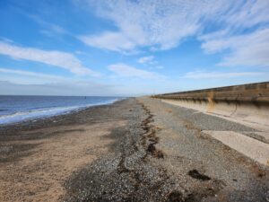 View of the tide line in front of a sea wall. Blue sky, Pebbles on the beach. Shoreline and sea wall meet at vanishing point in distance